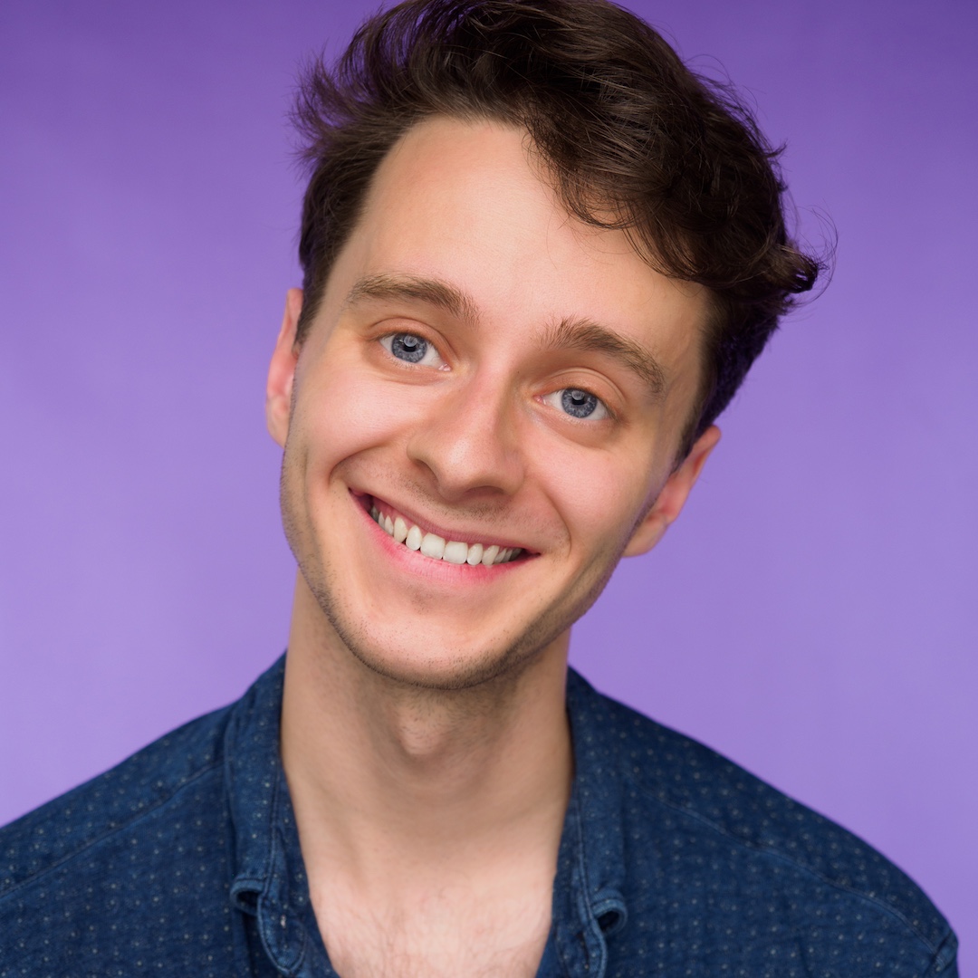 A photo of Liam, smiling, with short brown hair and an collared indigo shirt, against a light purple background.